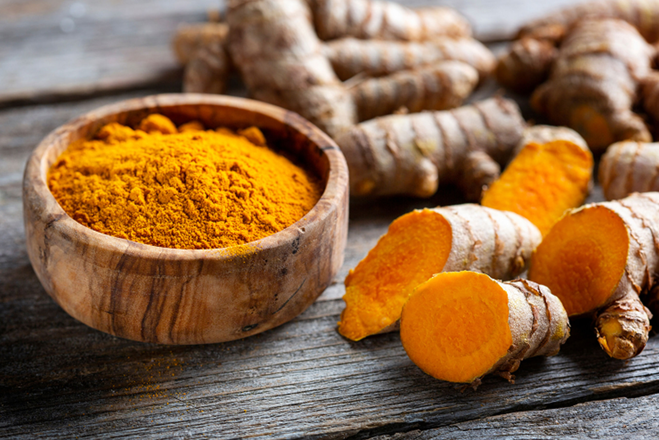 A wooden bowl filled with bright orange turmeric powder sits on a rustic wooden surface, surrounded by whole turmeric roots, some of which are sliced to reveal their vibrant yellow-orange interiors.