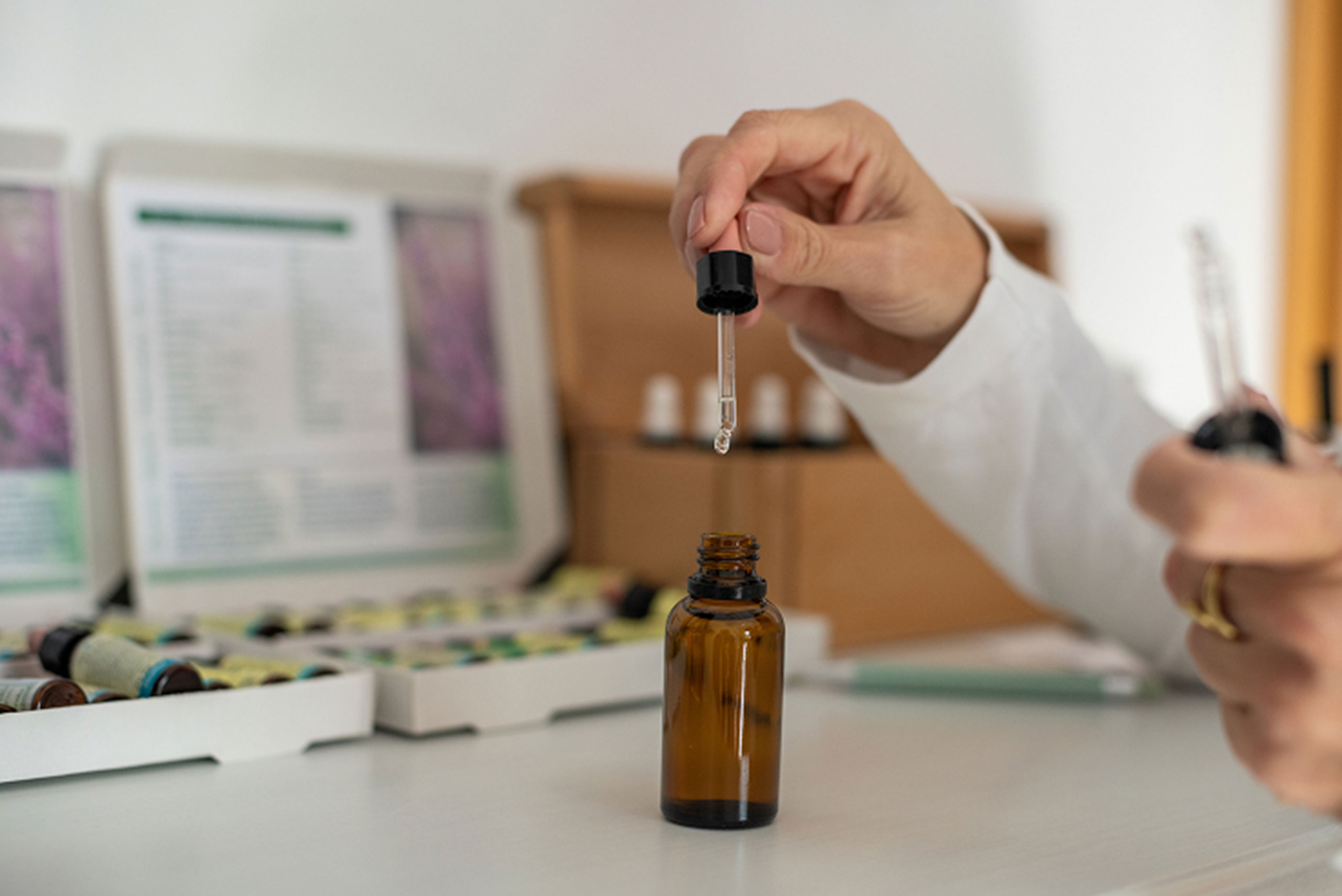 A person holds a glass dropper above an amber bottle, preparing to dispense liquid. In the background are boxes containing bottles and informational leaflets, suggesting a setting related to health or wellness.