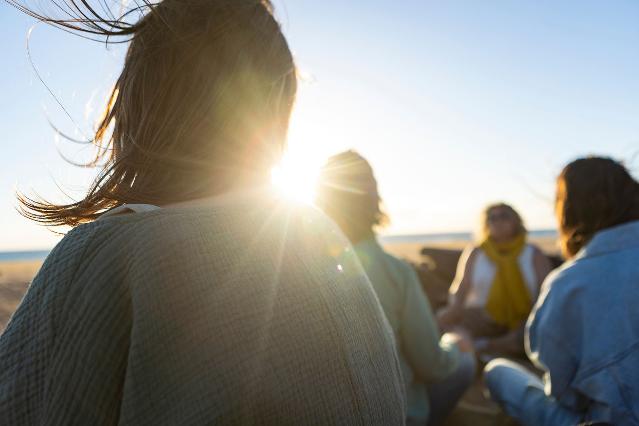 A group of people sit together on a beach at sunset, sunlight shining brightly behind them. Their faces are not clearly visible, and one person is in the foreground with windblown hair.