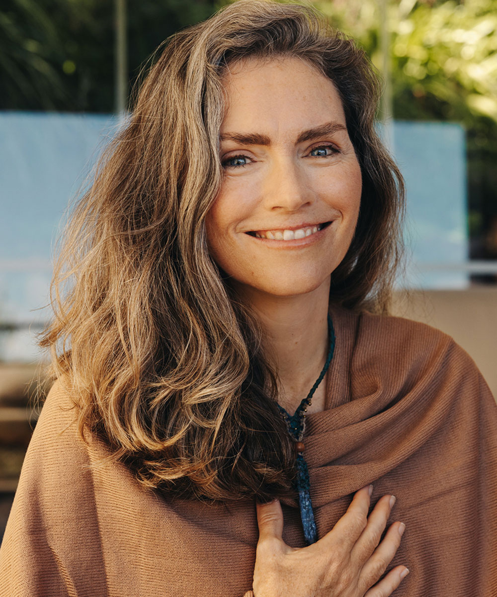 A woman with long wavy brown hair and blue eyes smiles gently, wearing a brown shawl and a necklace with a dark pendant, with one hand resting on her chest. The background is softly blurred with greenery.