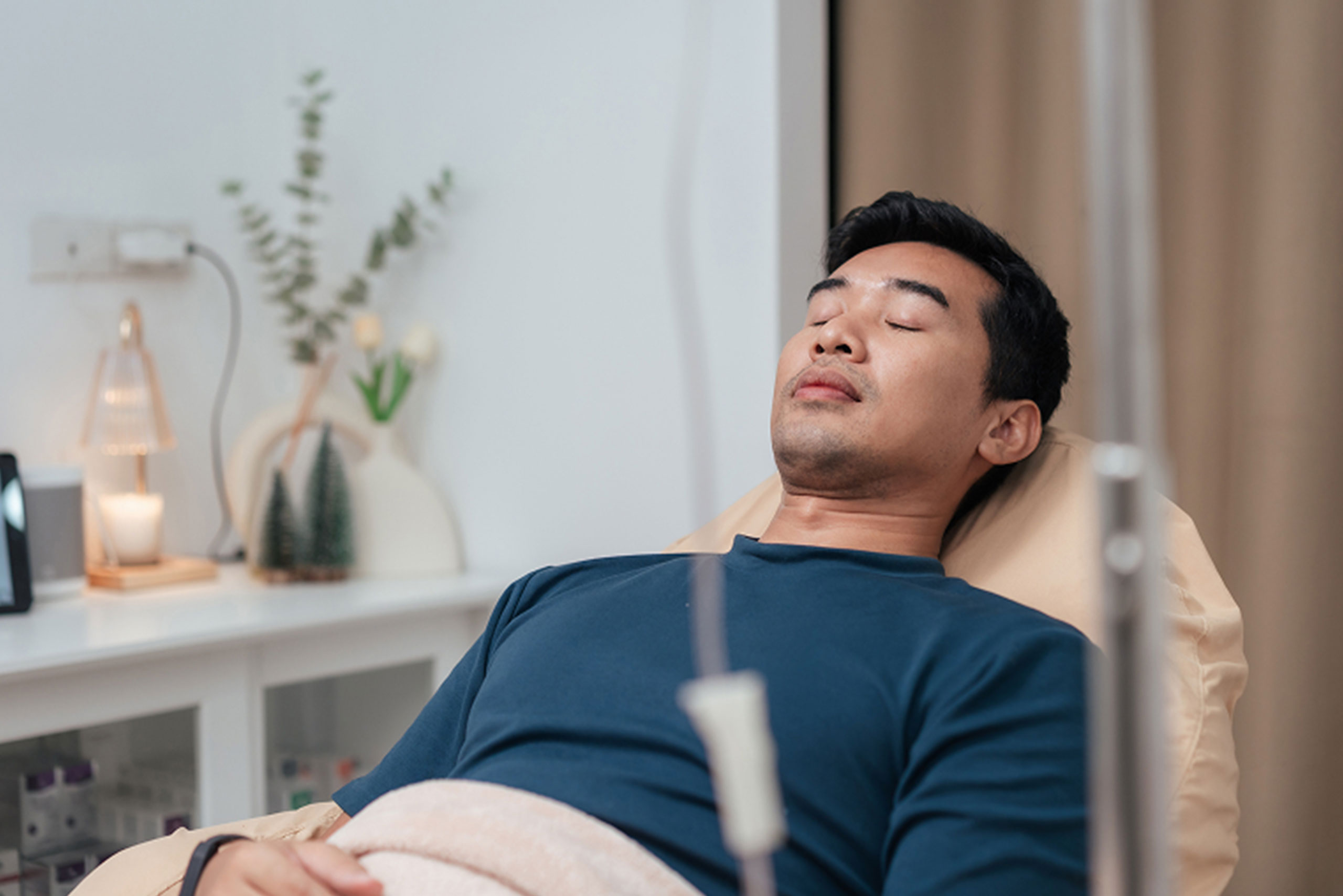 A man with dark hair lies on a recliner chair with his eyes closed, receiving an IV drip. The room appears calm, with soft lighting and decorative plants on a shelf in the background.