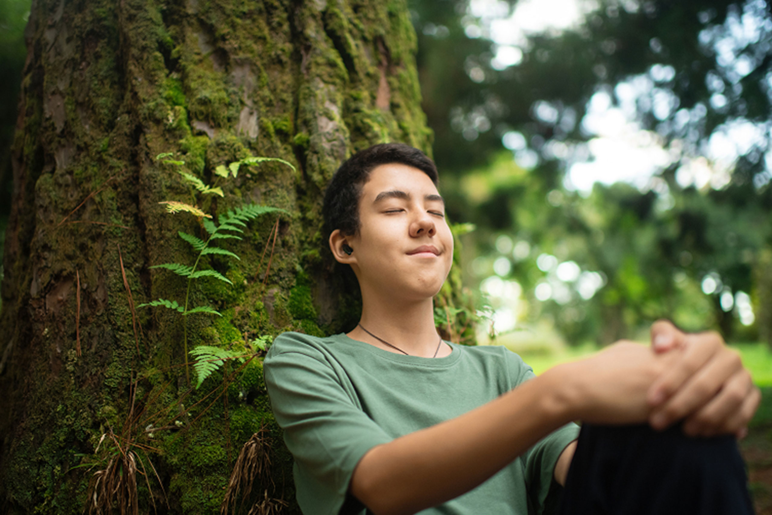 A young person sits with eyes closed, smiling peacefully while leaning against a mossy tree in a lush, green forest, enjoying a moment of relaxation in nature.