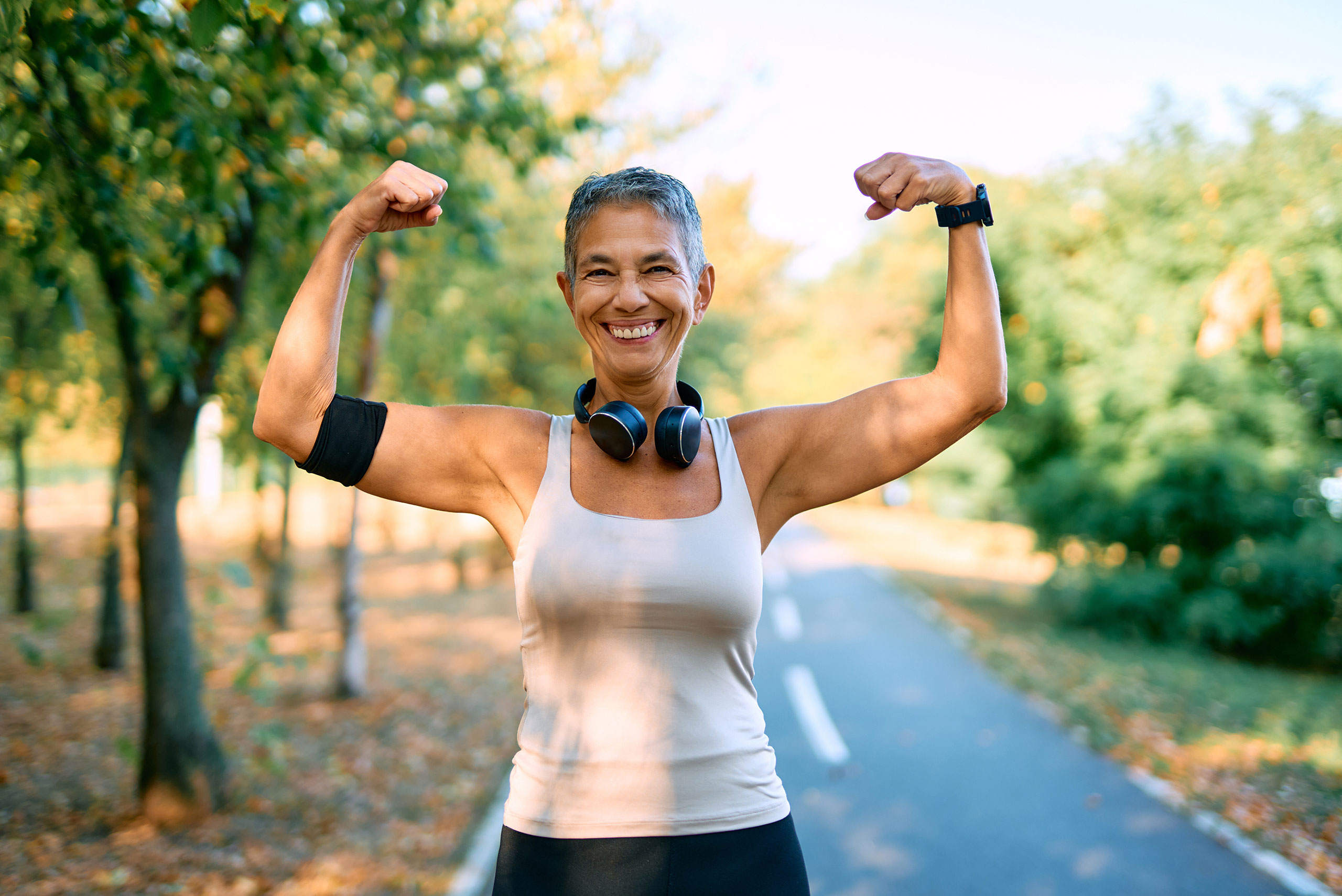 Smiling older woman in a tank top flexes her arms and shows her muscles while standing on a tree-lined outdoor path. She wears headphones around her neck and a fitness armband.