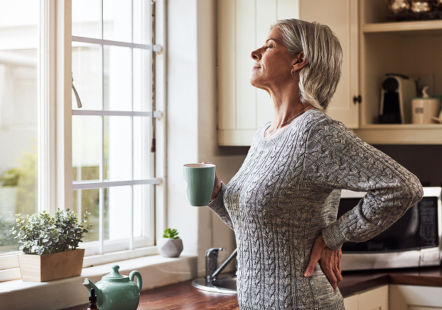 An older woman with gray hair stands in a kitchen, holding a mug and looking thoughtfully out the window. She wears a gray sweater, one hand on her back, reflecting on her journey with integrative cancer care Calgary offers.