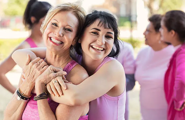 Two women in pink tops smile and hug each other outdoors, with other women in pink clothing chatting in the background. The group appears happy and supportive, possibly at a community or awareness event.