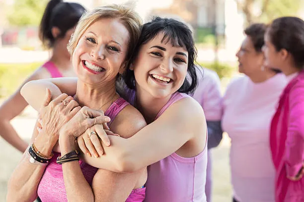 Two women wearing pink tops smile and hug each other outdoors, while a group of people in pink clothing stand and talk in the background, suggesting a supportive or awareness event.