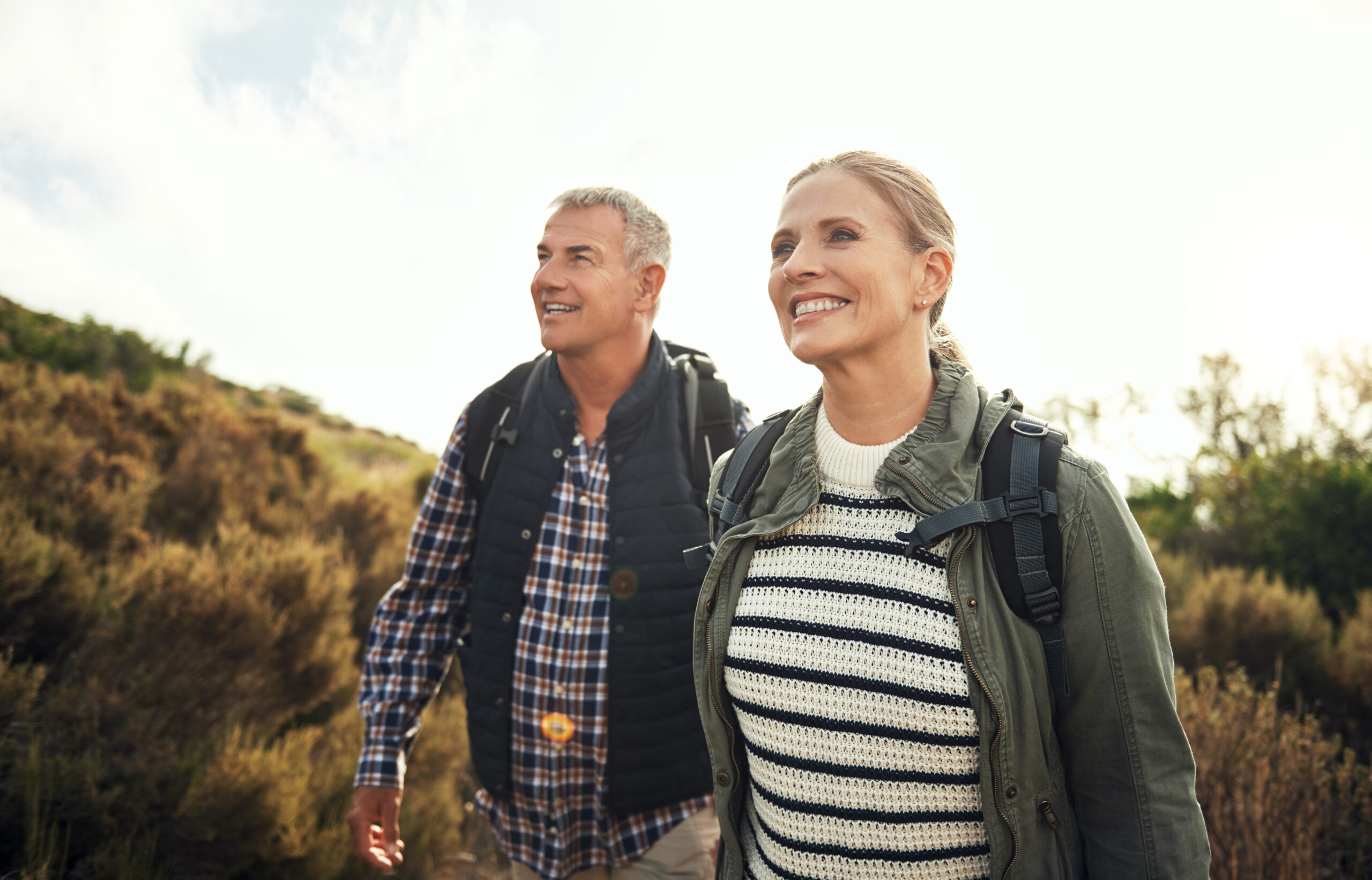 A smiling couple wearing casual outdoor clothing and backpacks walks along a nature trail on a sunny day, surrounded by green shrubs and a bright sky.