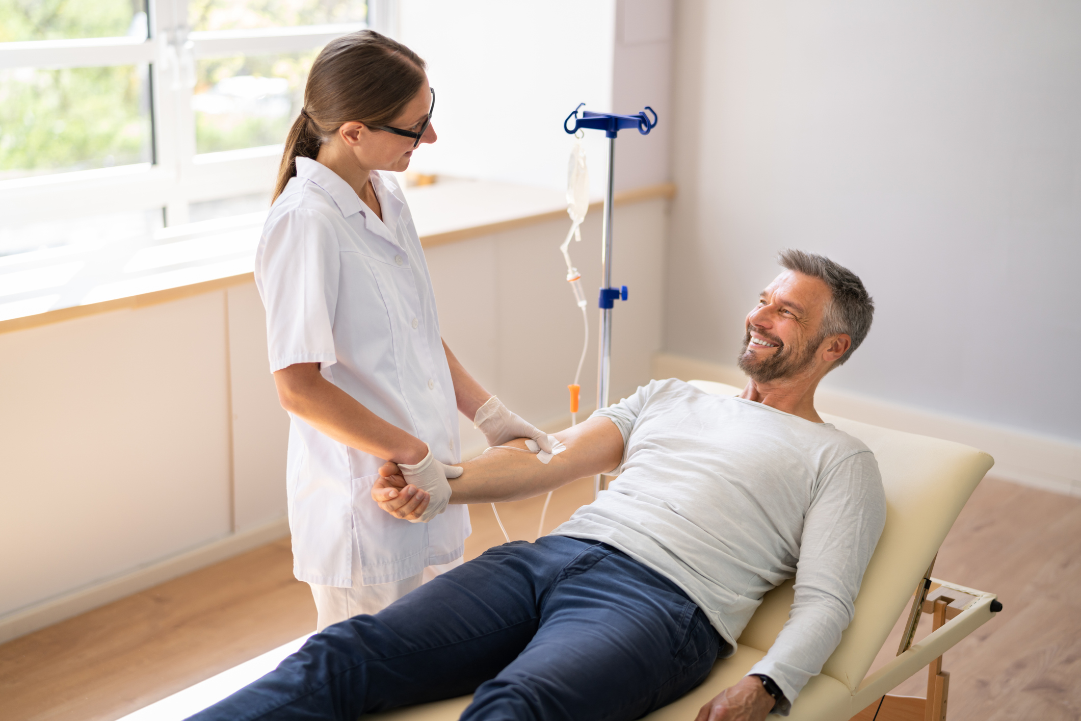 A healthcare professional prepares to administer an IV to a smiling man lying on a medical bed in a bright, clean clinic room.