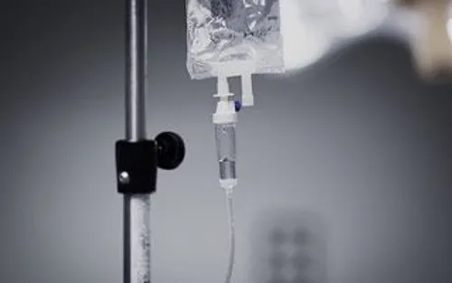 Close-up of an intravenous (IV) drip bag hanging on a metal stand, with tubing and a drip chamber visible, set against a blurred gray background.