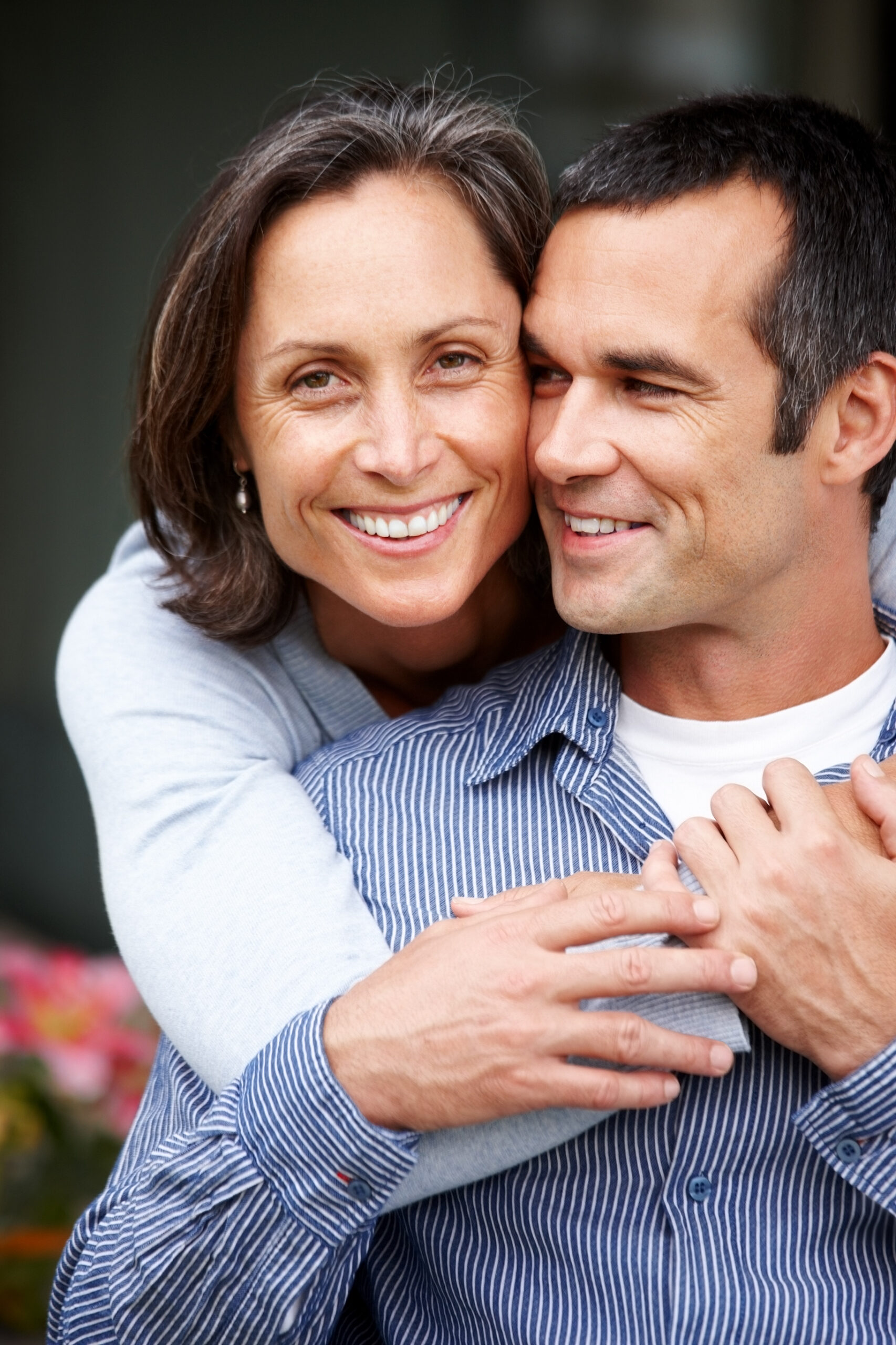 A woman with graying hair smiles and hugs a man from behind. Both appear happy and relaxed, with the man smiling and looking to the side. They are outdoors, and the background is softly blurred.